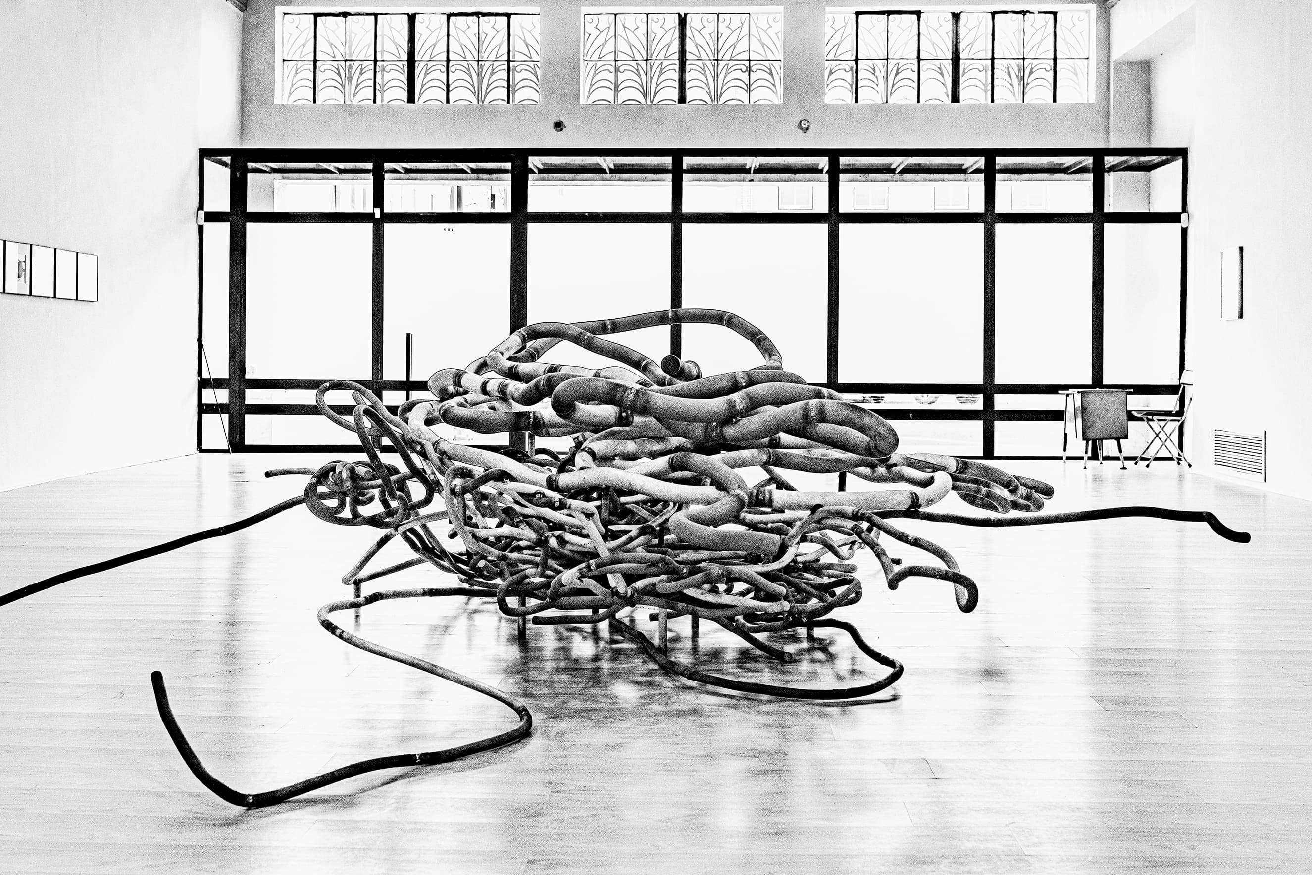 Photograph of a large mosaic artwork resembling a tangle of lines. It is hanging on a wall inside of an unfinished space. The artwork is flanked by large concrete columns and the space is strewn with wires, pipes and construction materials.