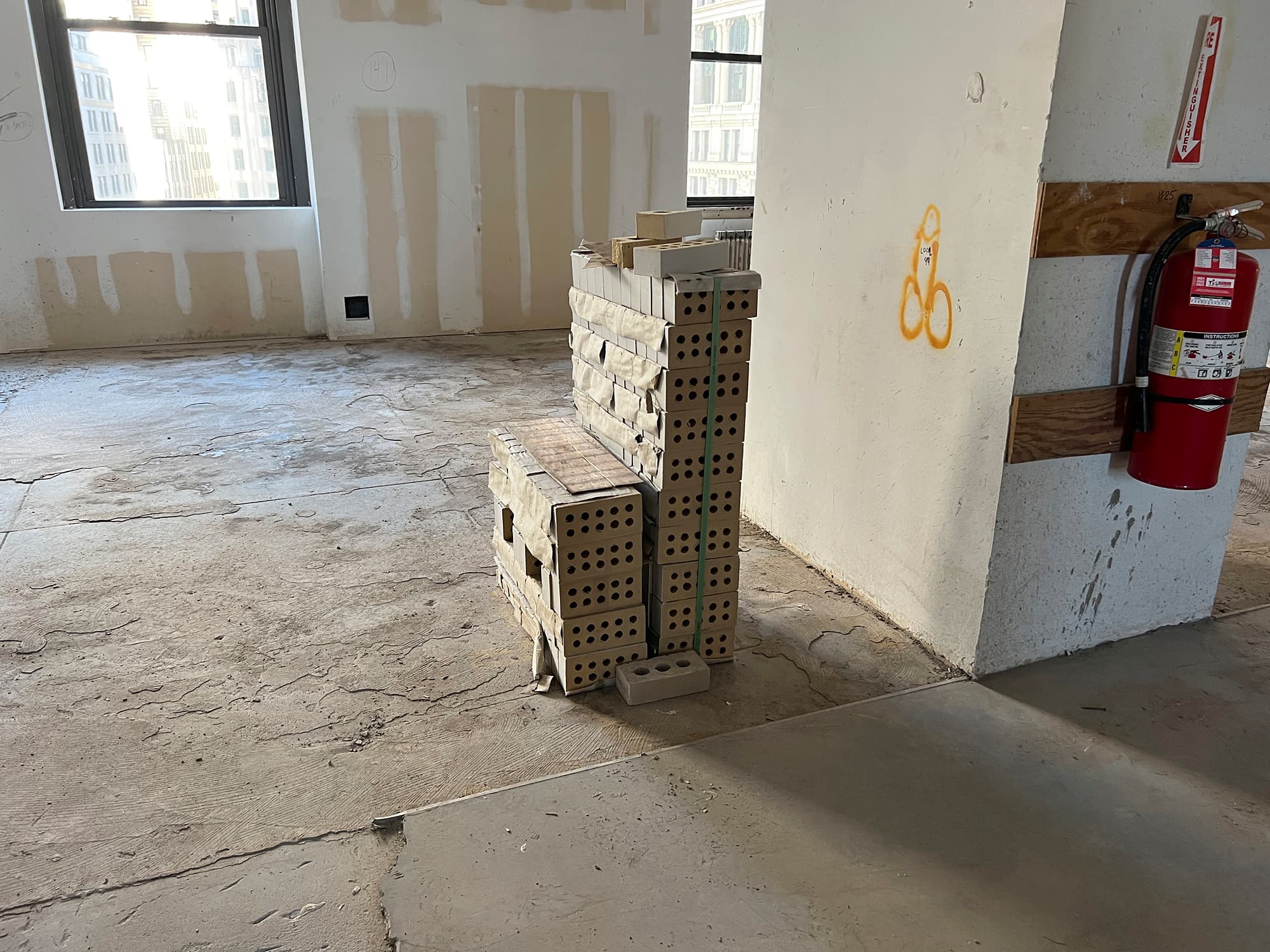 A color image of a raw interior space with graffiti on the walls and a fire extinguisher affixed to a column on the right. At center, a stack of tan bricks, still in construction strapping, sits on the floor.