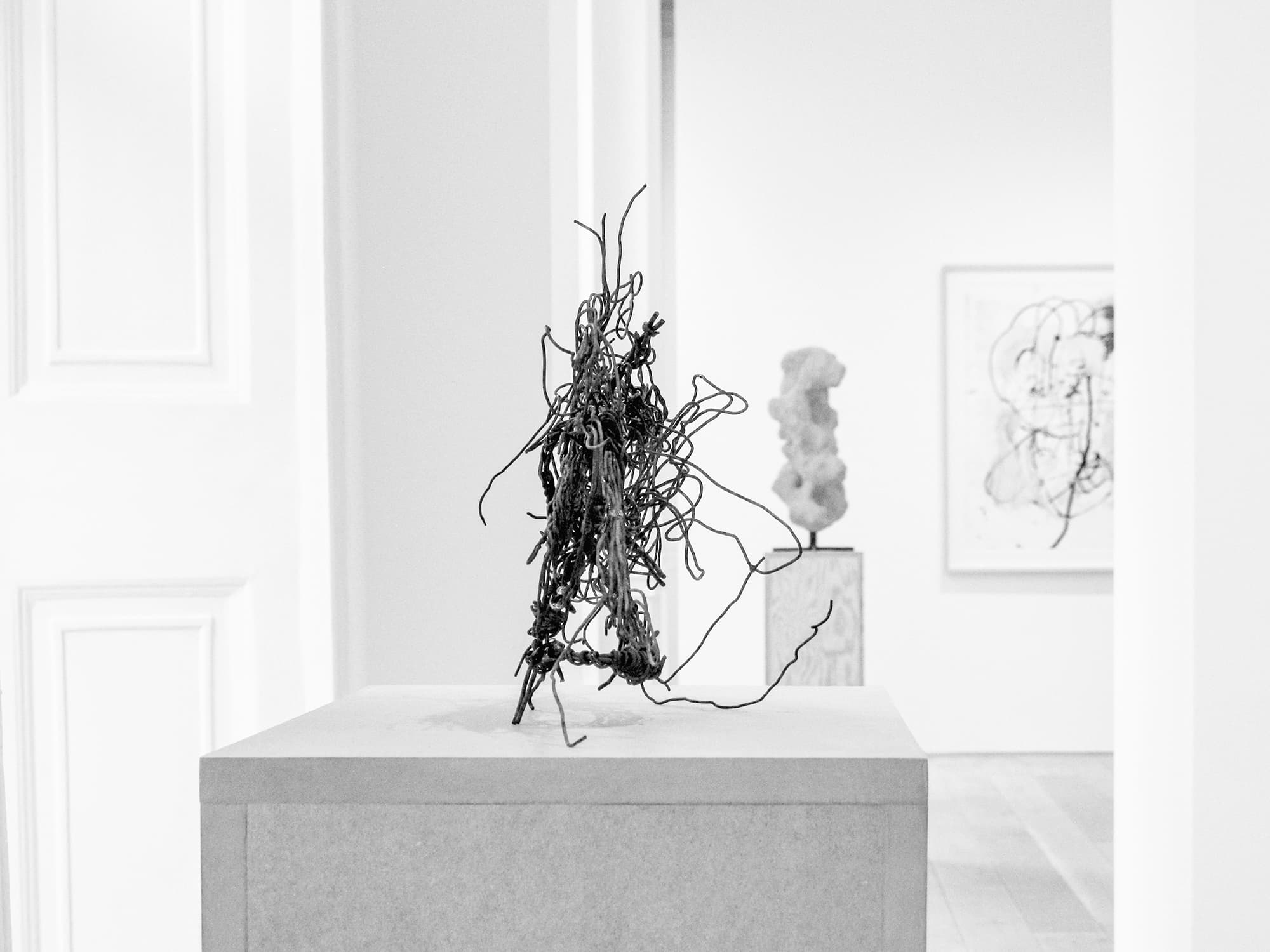 A black and white photograph of a small wire sculpture on a pedestal in a gallery context. The wire is densely bent together in irregular formations; the entirety stands on a few of the wires conjuring a figure-like shape. In the background, another room houses a clay-like sculpture on a pedestal and a portion of a framed piece of art hanging on the wall.