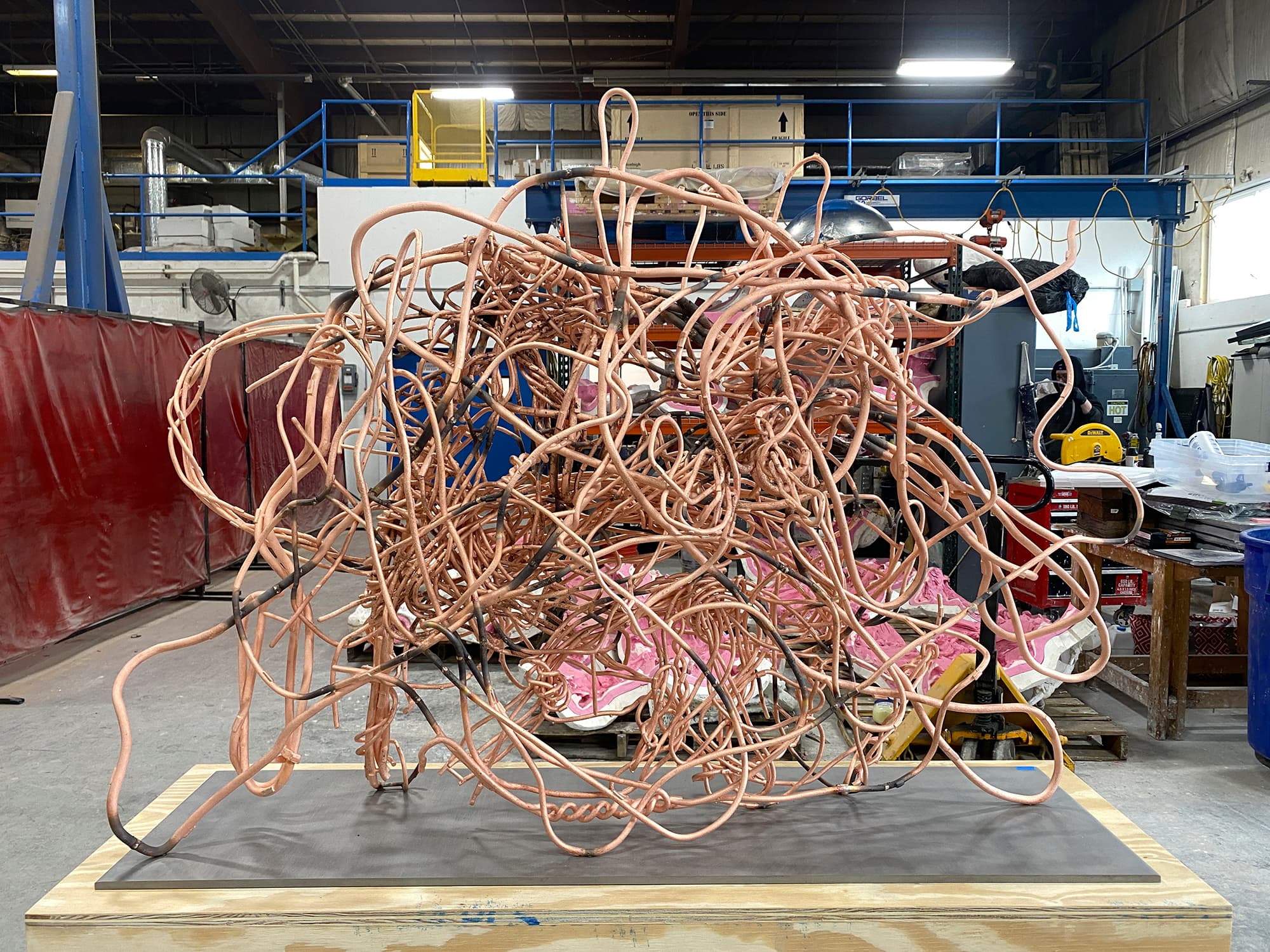 A color image of a copper sculpture attached to a plinth in an industrial space with welding equipment in the background and fluorescent lights overhead. The sculpture is made of thin copper-toned rod and is twisted into a tight conglomeration.