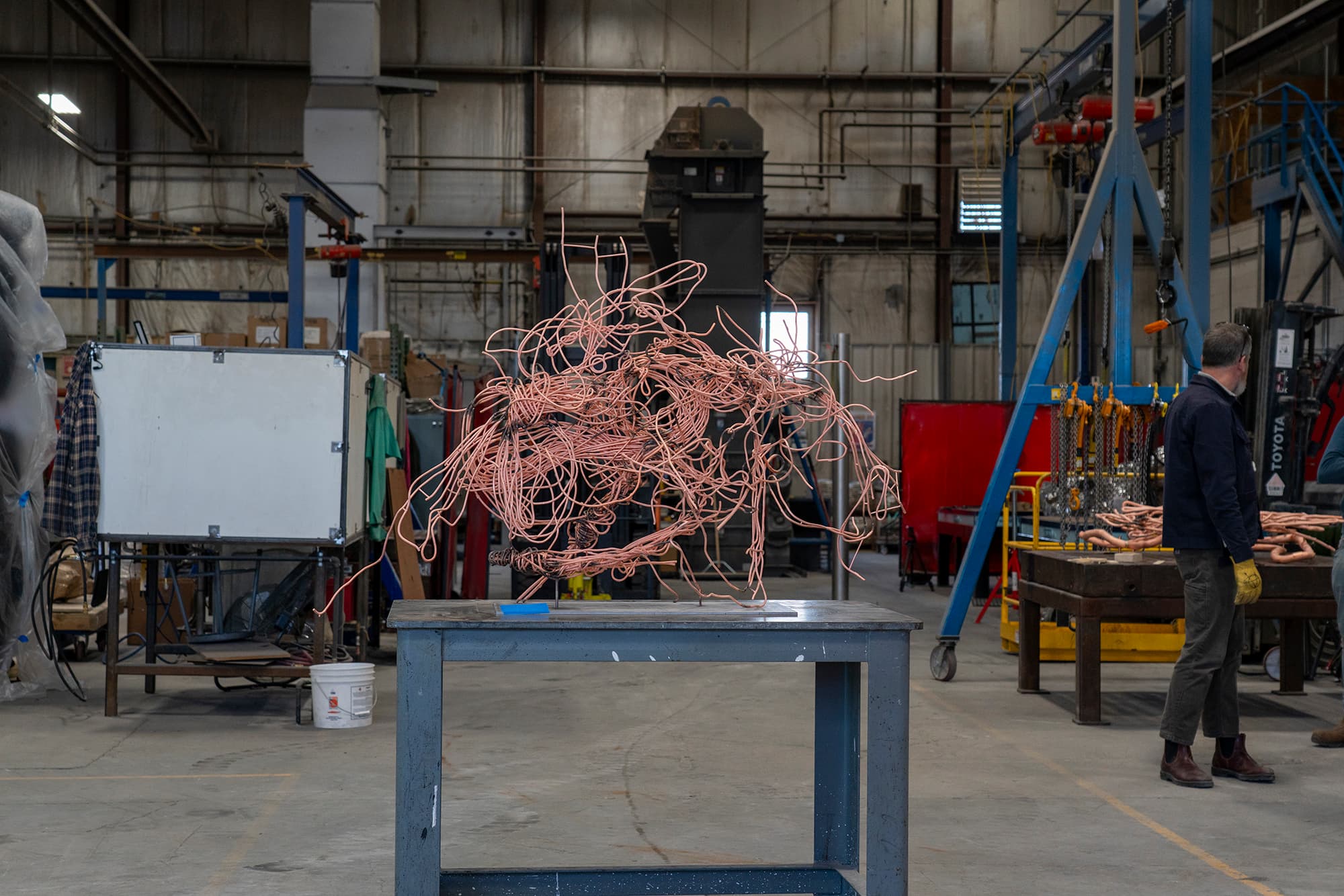 A color image of a copper rod sculpture on a blue work table in an industrial space. Welding equipment fills the background and a person stands at the right edge of the image, turned toward the rear of the space.