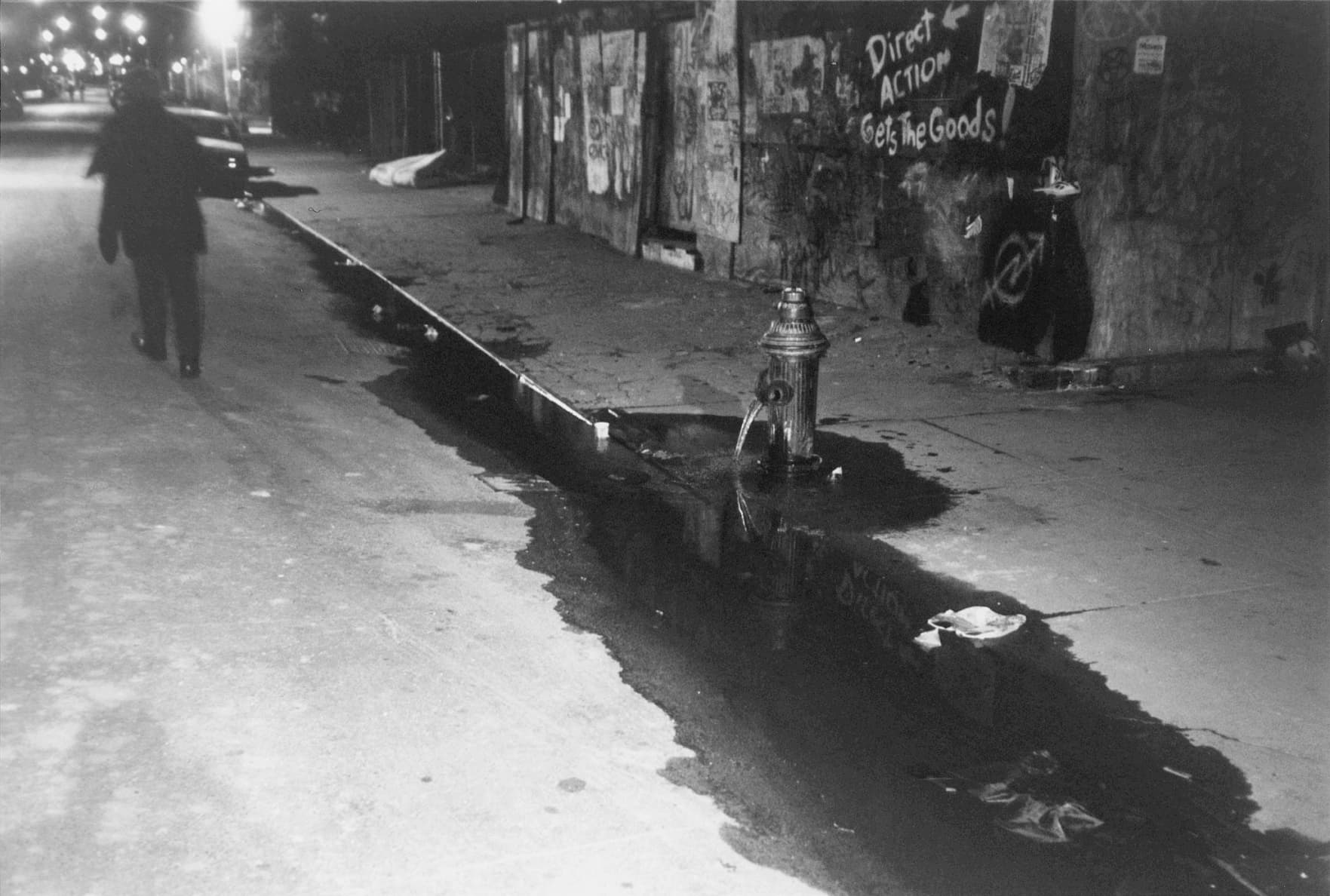 A black and white photograph that appears aged in which a silhouetted figure walks down an urban street at night lit by street lamps. In the foreground, a large black puddle cuts across the image, creating a diagonal oblong shape.