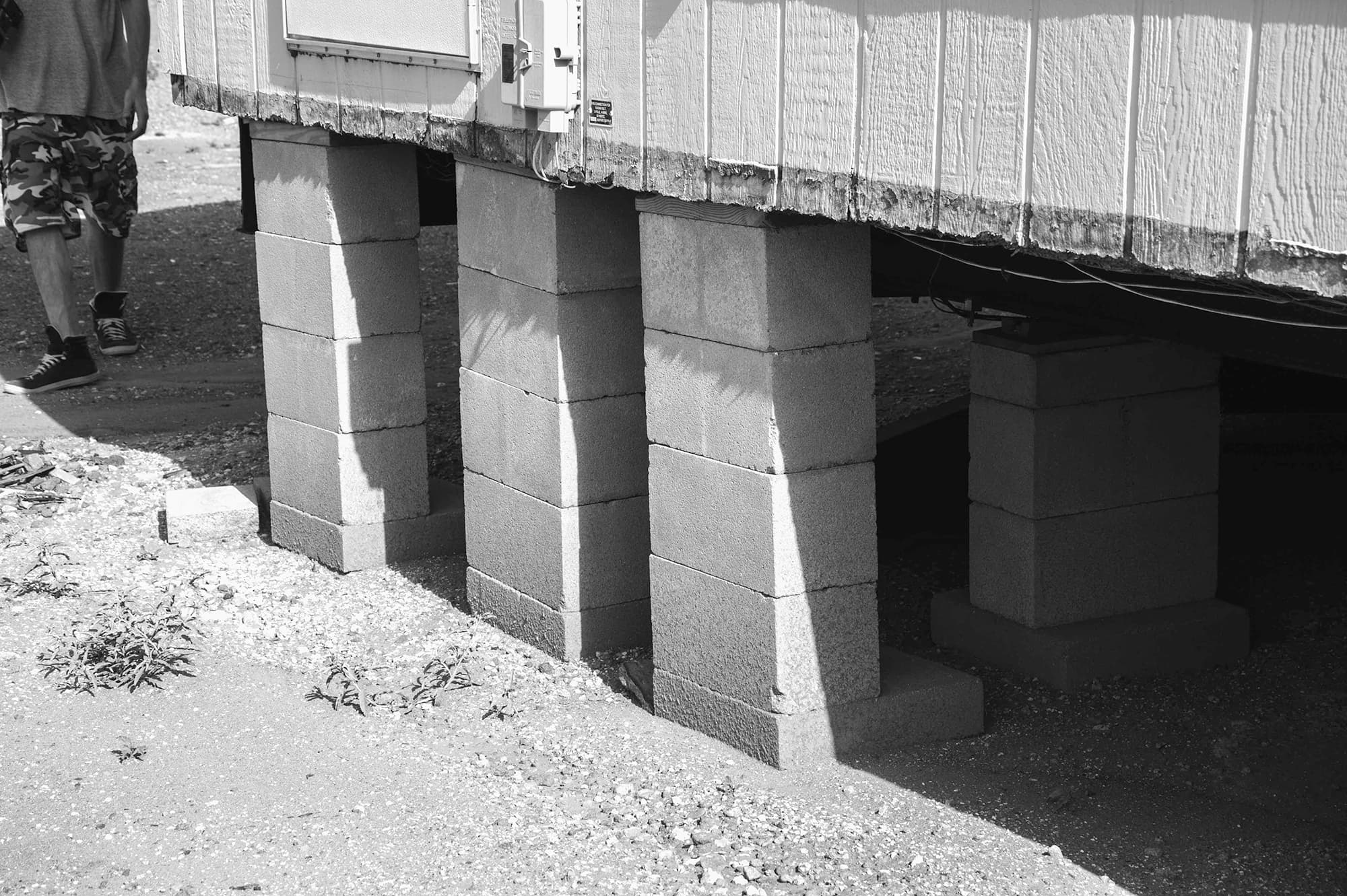 A black and white photograph of stacks of concrete blocks supporting a structure. The sharp sunlight creates a pattern of shadows and textures. At the top left of the image the lower portion of a shadowed figure in sneakers and shorts, is captured midstep.