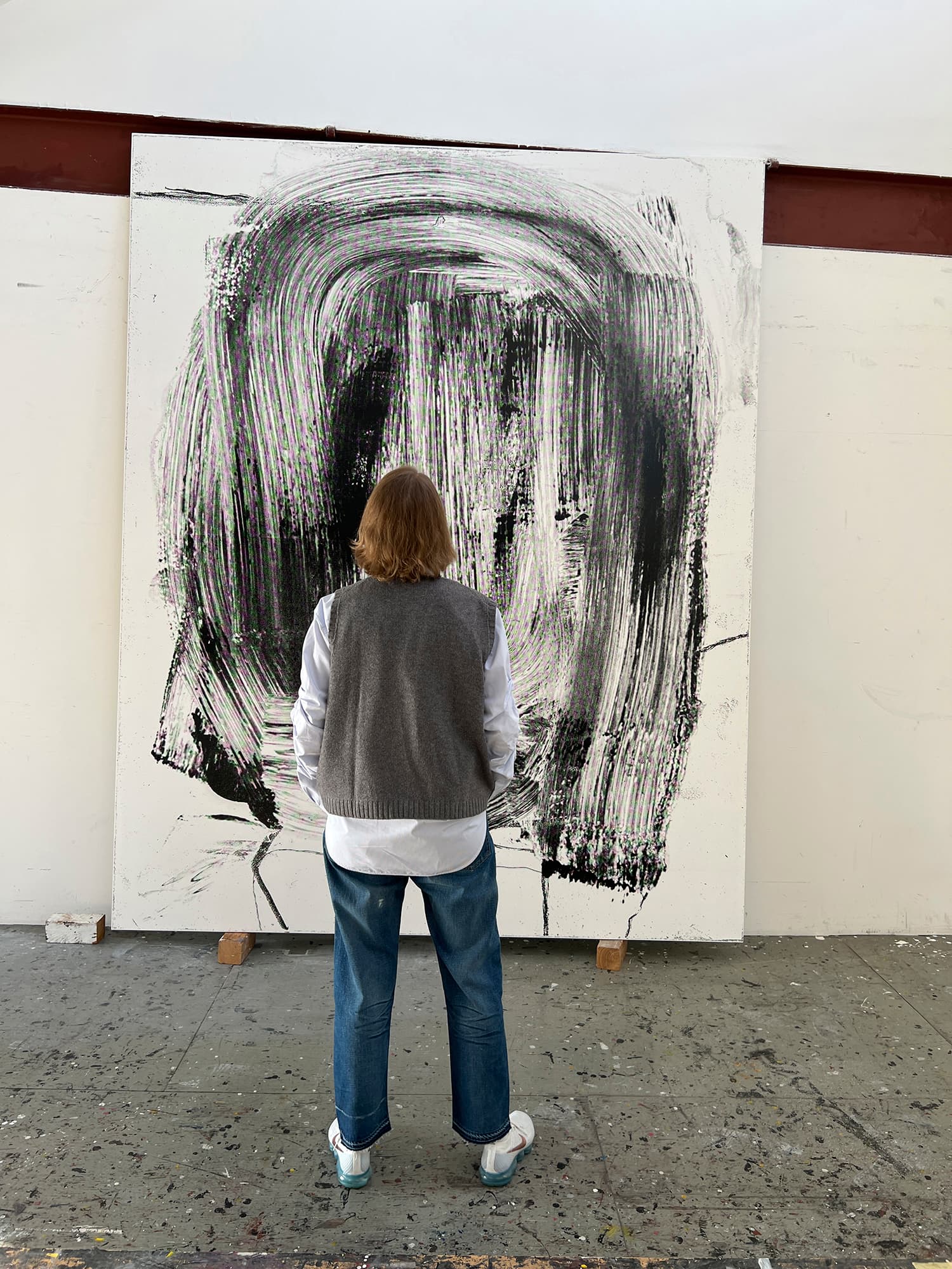 A color image of a person standing in front of a painting which leans on a wall. The person’s back faces the camera. The painting appears to be enlarged brushwork, black pigment on a white support.