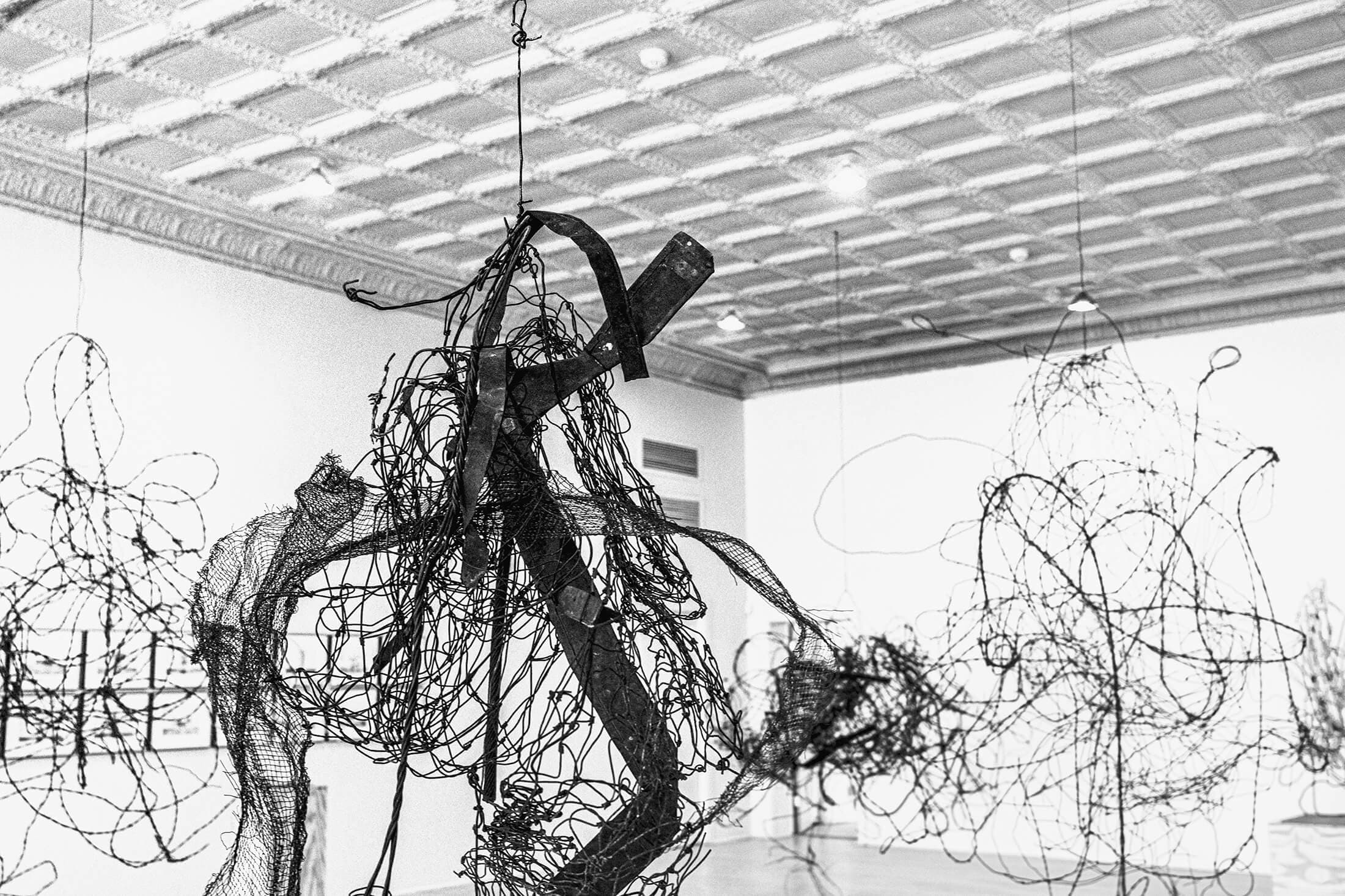 Photograph of a large mosaic artwork resembling a tangle of lines. It is hanging on a wall inside of an unfinished space. The artwork is flanked by large concrete columns and the space is strewn with wires, pipes and construction materials.